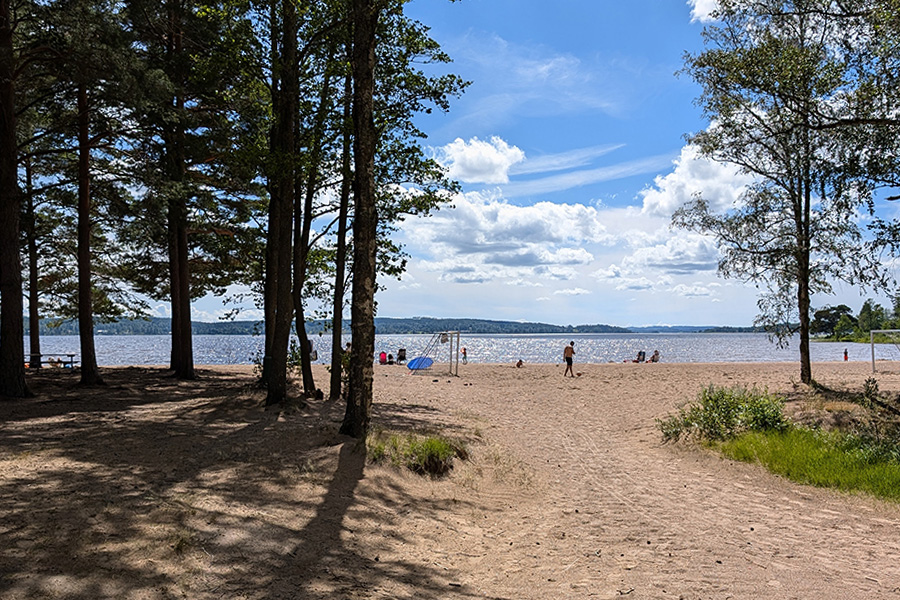 På bilden syns en sandstrand omgiven av träd som skapar en naturlig skugga. Stranden leder till en sjö eller ett hav med glittrande vatten under en klarblå himmel med några vita moln. I bakgrunden kan man se människor som badar och kopplar av på stranden. Några av dem sitter under parasoller och några är ute och promenerar. Det finns också två fotbollsmål placerade på stranden.