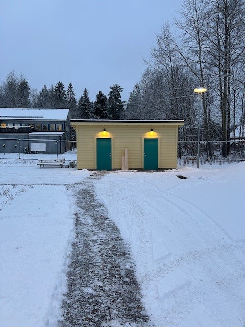 En gul förrådsbyggnad med två gröna dörrar, två lampor lyser ovanför. Marken är täckt med snö.