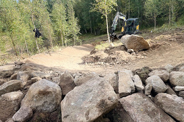 En grävmaskin i ett skogsområde med större stenar i förgrunden. 