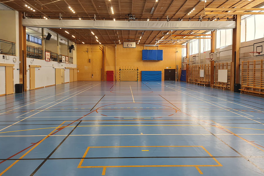 Idrottssal med blå golvbeläggning. Det finns basketkorg, handbollsmål och tjockmattor. I taket finns en skiljevägg. Längs ena väggen finns bänkar och ribbstolar. 
