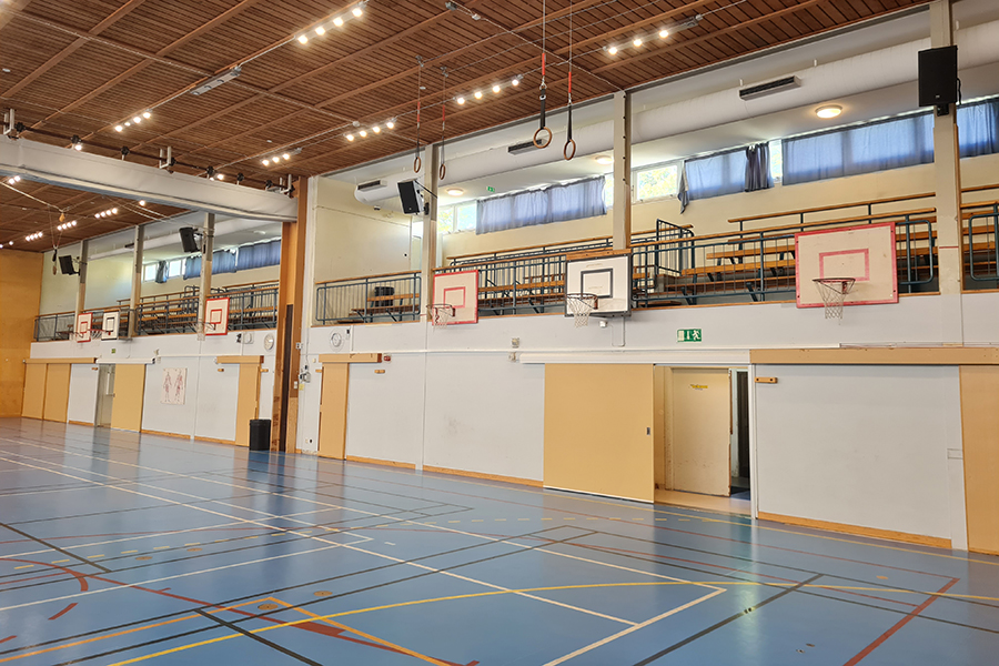 Idrottssal med blå golvbeläggning. Bilden visar ena långväggen där det finns basketkorgar och läktare.