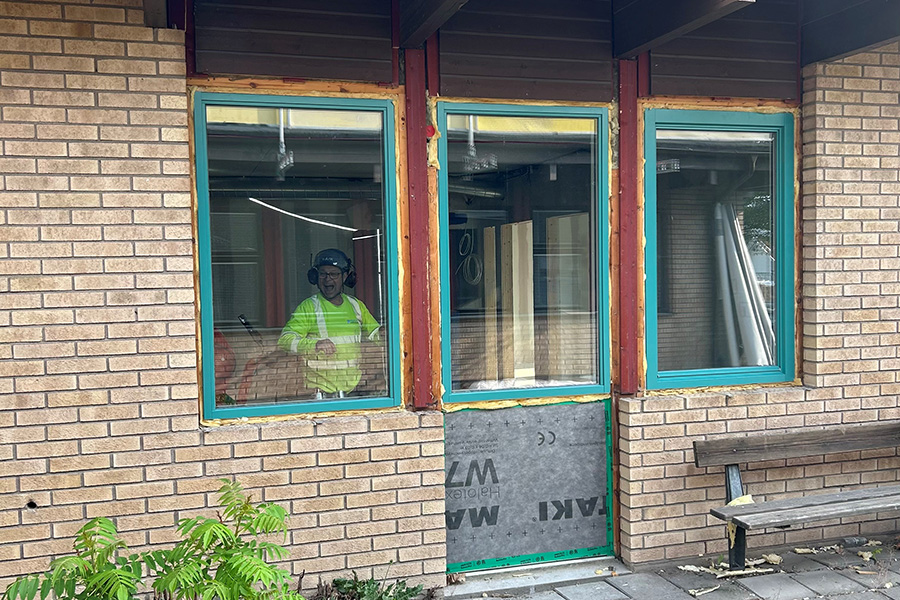 En bild på en byggnad men en gulaktig tegelvägg med gröna fönsterkarmar. I ena fönstret står en person i arbetskläder och tittar ut.