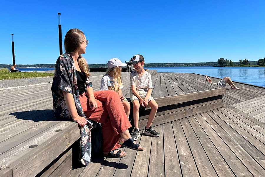 En mamma och två barn sitter tillsammans på en brygga och skrattar.