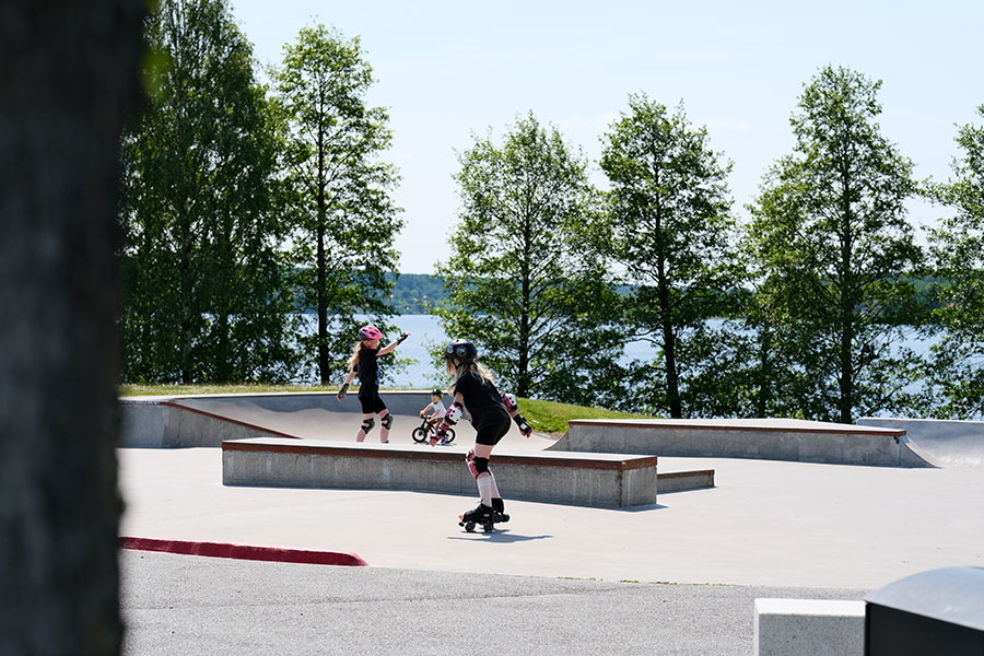 Två barn åker inlines i en skatepark tillsammans. I bakgrunden kan man skymta en sjö.