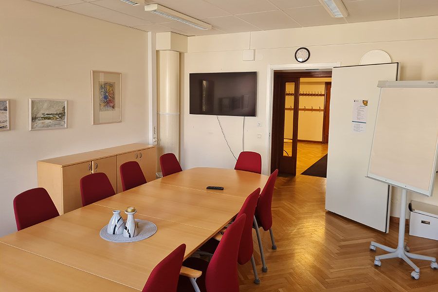 Mötesrum med bord, nio röda stolar. Hurts och tavlor på en vägg. Digital skärm och dörr längs en vägg. Fristående whiteboard-tavla på hjul. 