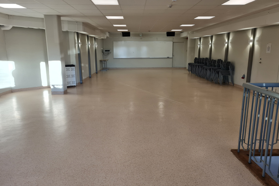 Stor möteslokal, tom golvyta, stort antal stolar efter en vägg. Längst fram en Whiteboardtavla och två högtalare i taket.