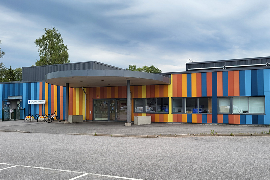 Mångfärgad byggnad med en stort tak ovanför entré. Asfalterad yta framför.