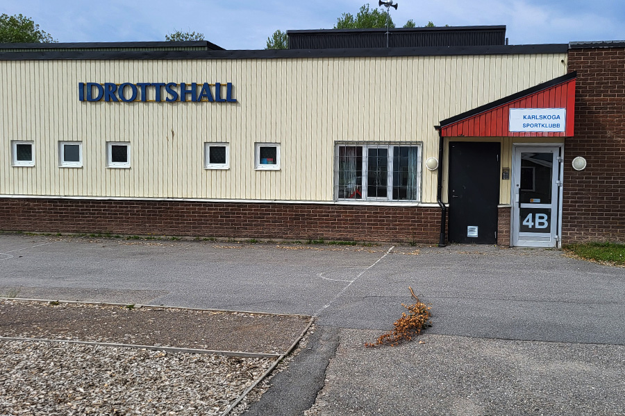 Skolbyggnad med ljus fasad och rött tegel. Entrédörr till idrottshallen. Asfalterad yta framför.