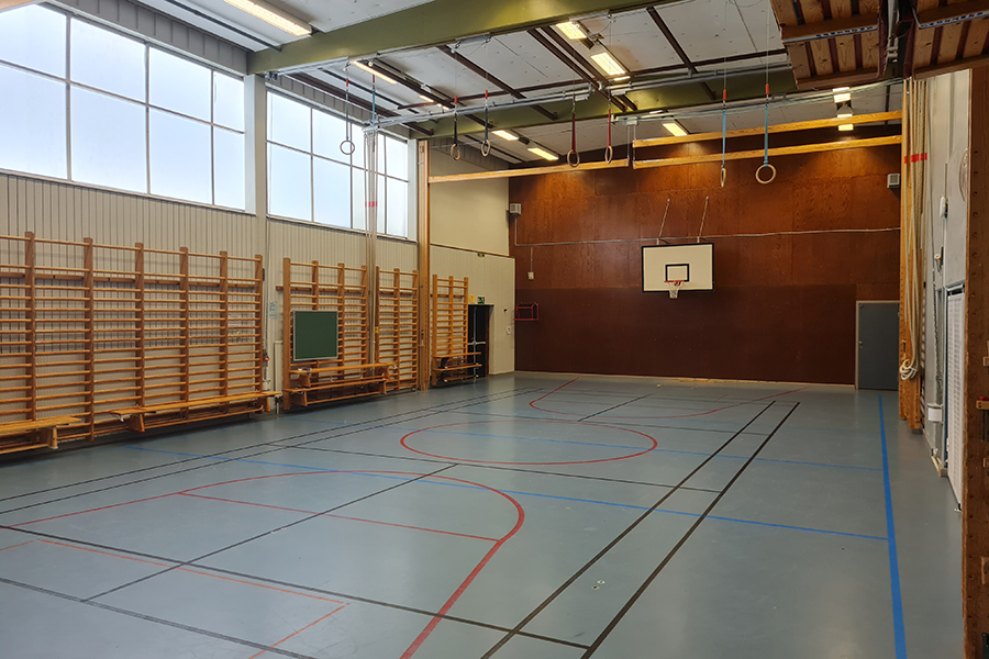 Idrottssal med grå golvbeläggning. Basketkorgar på kortvägg, redskap i taket. Ribbstolar och bänkar längs ena väggen. Dörr och nödutgång på kortsidan.