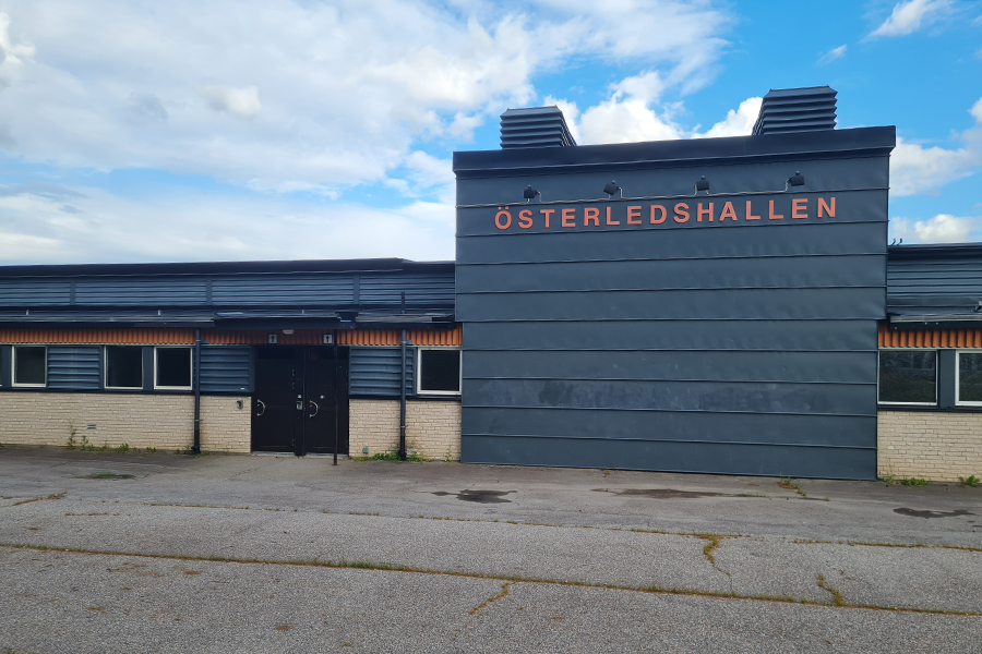 Entrédörrar till skolbyggnad med fasad i vitt tegel och blått trä. Stor skylt med text Öserledshallen. Asfalterad yta framför.