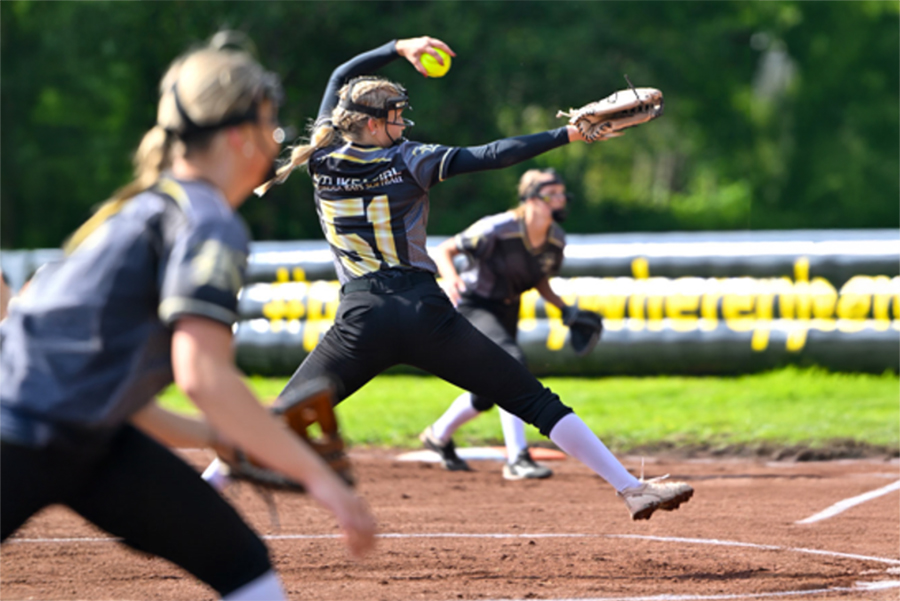 En softbollsspelare i aktion, klädd i en svart och gul uniform med texten "Karlskoga Bats" på ryggen. I förgrunden är en spelare, som står på en mjuk grusplan, är i en pitching-position och har precis släppt en gul softboll. I bakgrunden syns ytterligare lagkamrater i liknande uniformer, och landskapet är omgivet av gröna träd.