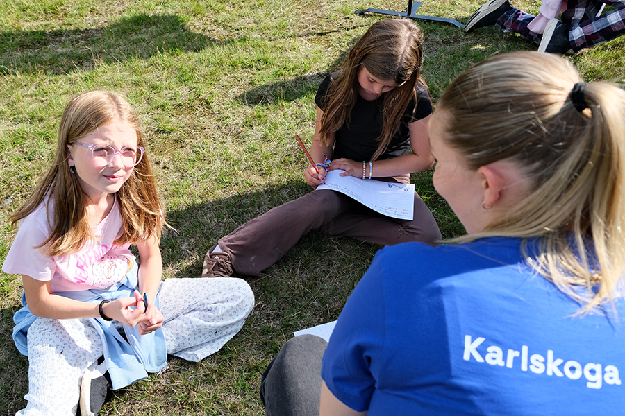 Tre barn sitter på gräset och skriver. En tröja visar texten ”Karlskoga”.