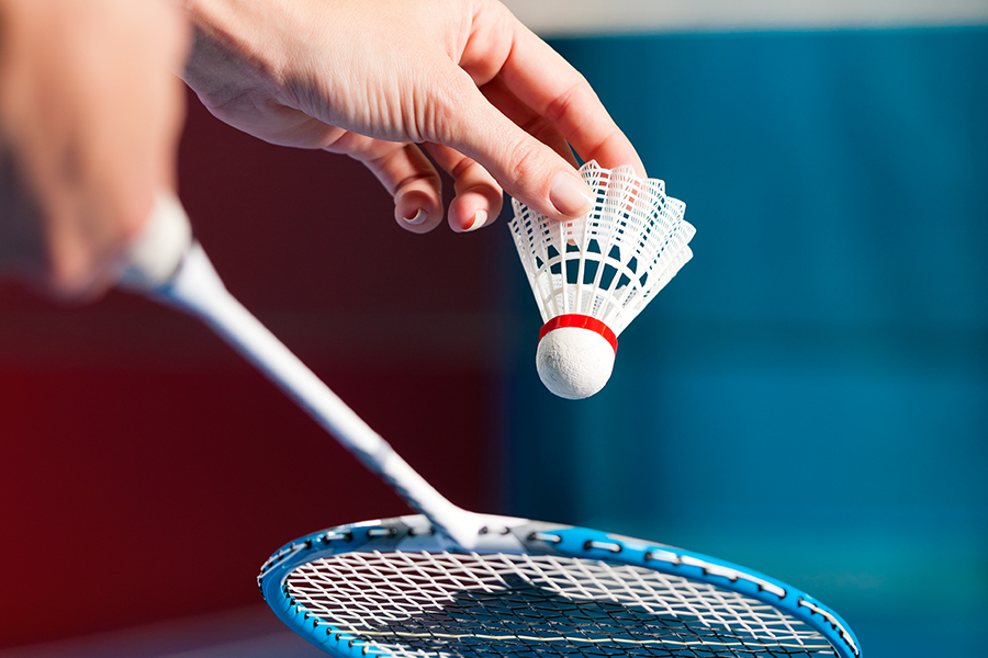 En hand håller en badmintonfjäderboll. En blå och vit racket syns i förgrunden.
