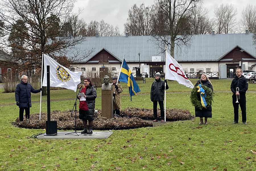 Sex personer står på en. Två håller flaggor i handen och en pratar i en mikrofon.