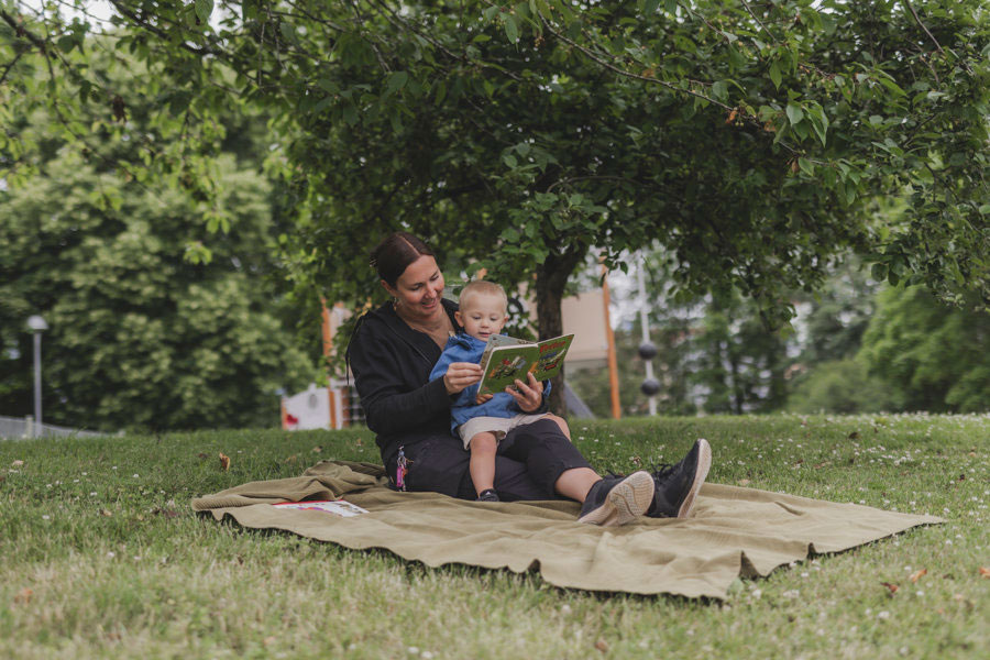 En kvinna som sitter på en filt ute i naturen och läser en bok för ett barn.