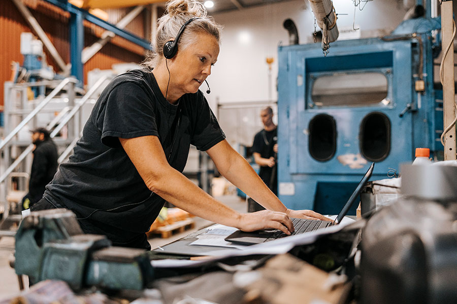 Äldre kvinnlig lagerarbetare med headset som använder en bärbar dator i en tillverkningsfabrik.