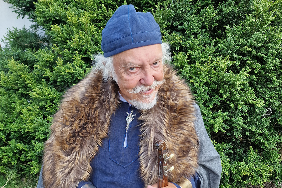 En man med grått hår och skägg och en ooälskrage runt halsen och en blå mössa på huv