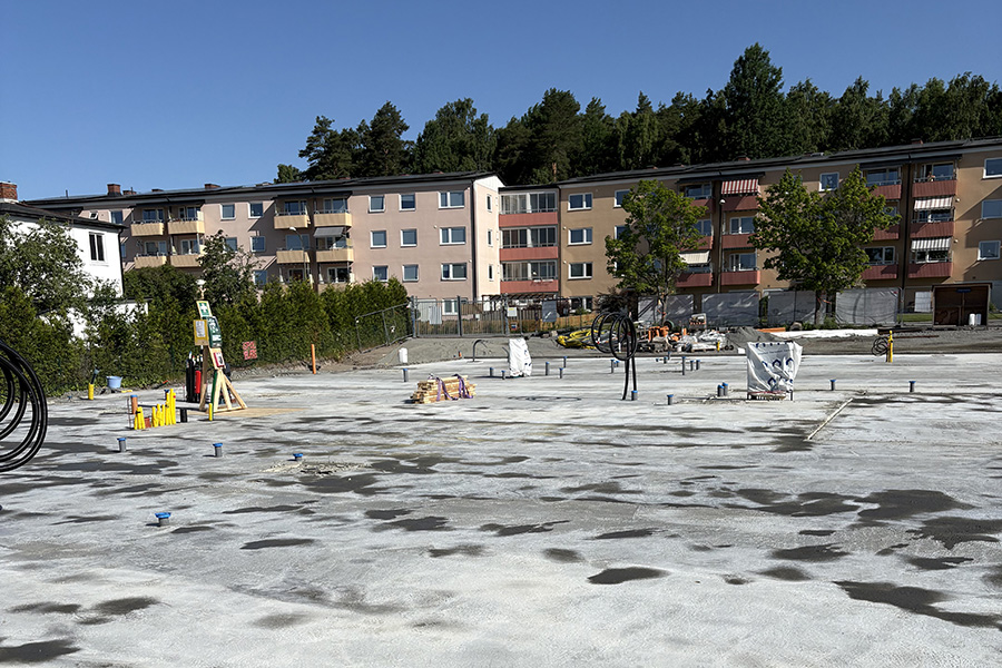 En byggarbetsplats med betongplatta och byggmaterial. Lägenhetskomplex med balkonger syns i bakgrunden, omgivet av gröna träd.