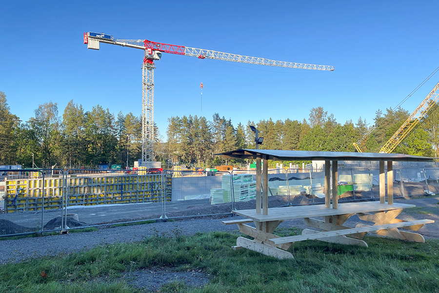 En träbänk som står framför en byggarbetsplats. I bakgrunden syns en kran. 