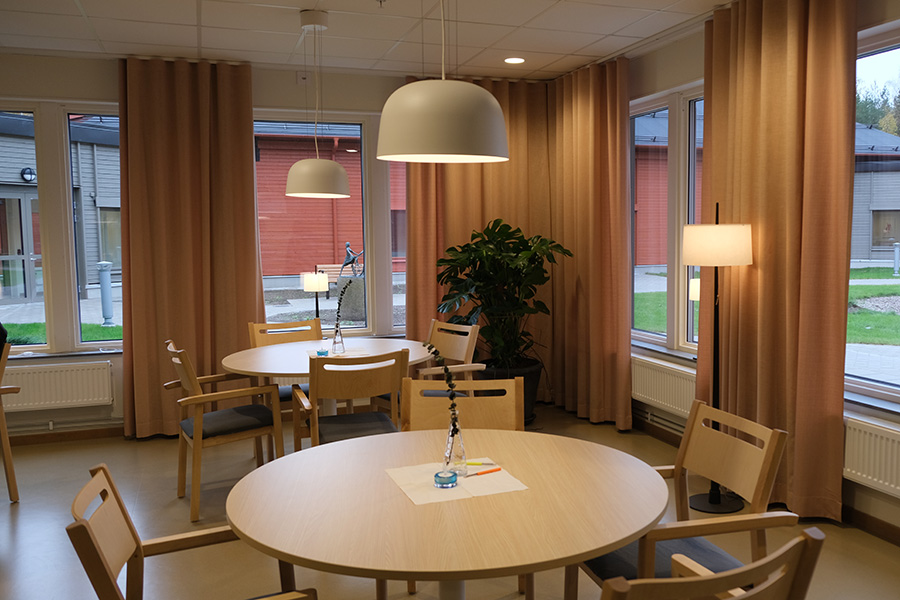 Ett rum med flera runda bord och tillhörande stolar. I bakgrunden syns fönster med gardiner.