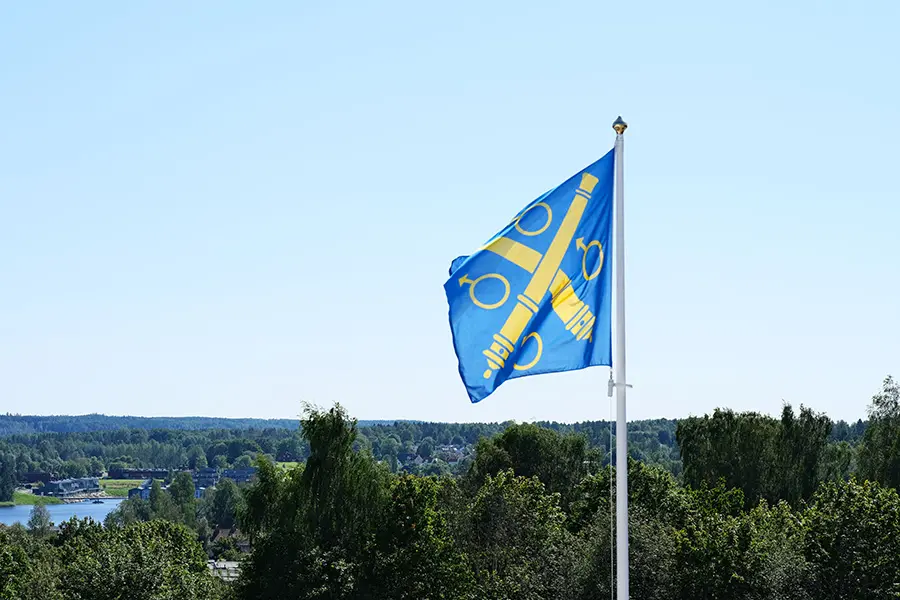 En blå flagga med Karlskoga kommuns kommunvapen i gult vajar mot en ljusblå himmel. I fjärran finns skog och en sjö.