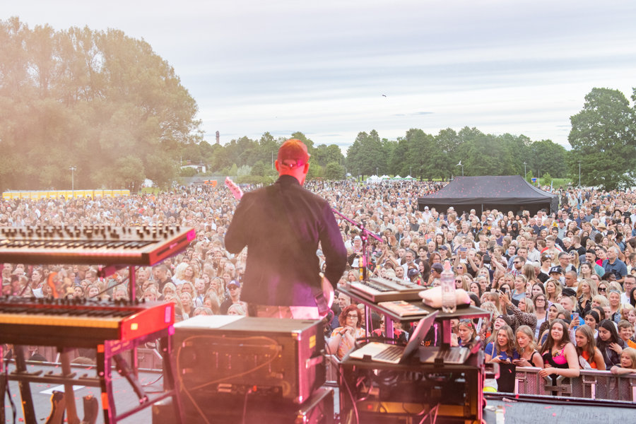 En man i keps står på en stor scen och håller en gitarr. Bakgrunden utgörs av ett stort publikhav och grönskande träd.