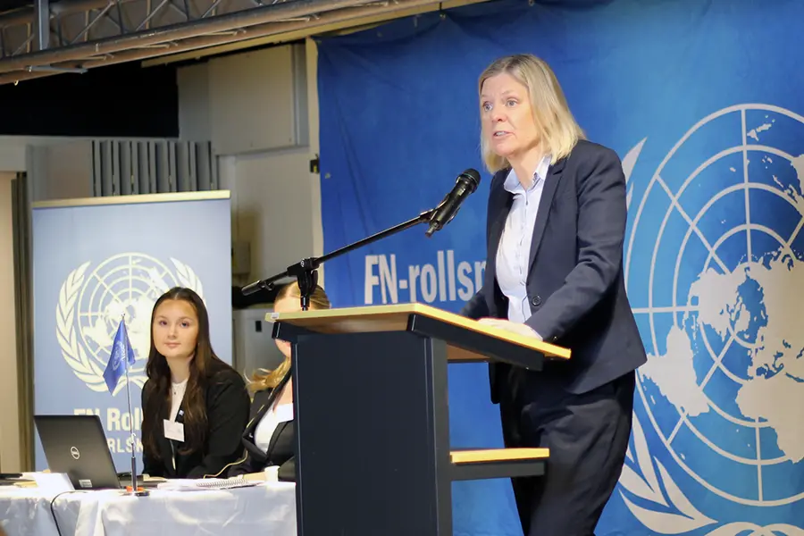 Socialdemokraternas partiledare Magdalena Andersson står i en talarstol och pratar in i en mikrofon. Hon bär mörkblå kostym och en ljusblå skjorta. I bakgrunden sitter två kvinnliga elever vid ett bord och bakgrunden pryds av FN:s blåvita logga föreställande en inringad världskarta. 