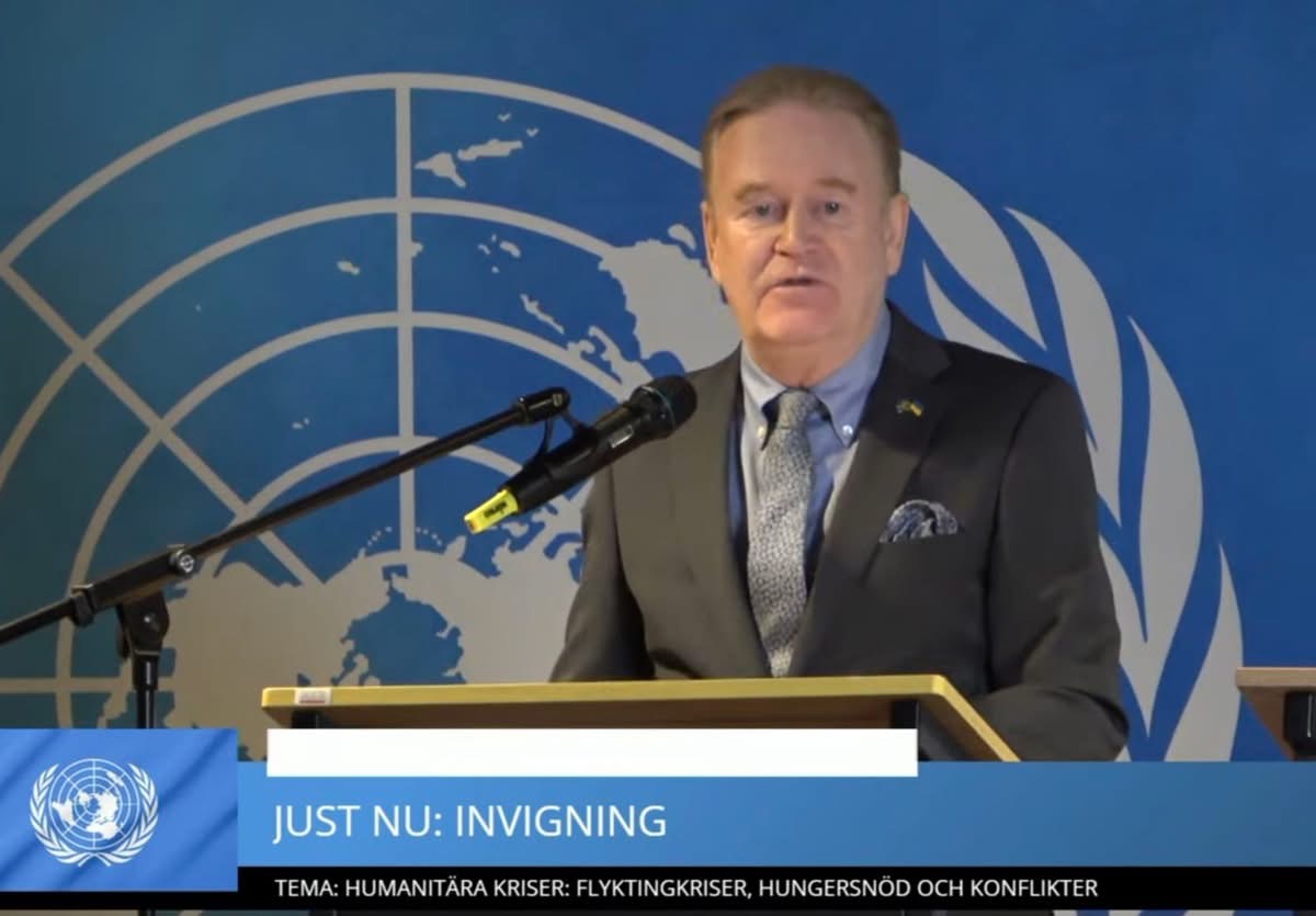 En man klädd i kostym och slips står i en talarstol och pratar in i en mikrofon. I bakgrunden är FN:s blå-vita logotyp och längst ned i bild syns ett tillagt blått fält där det framgår att man just nu inviger evenemanget.