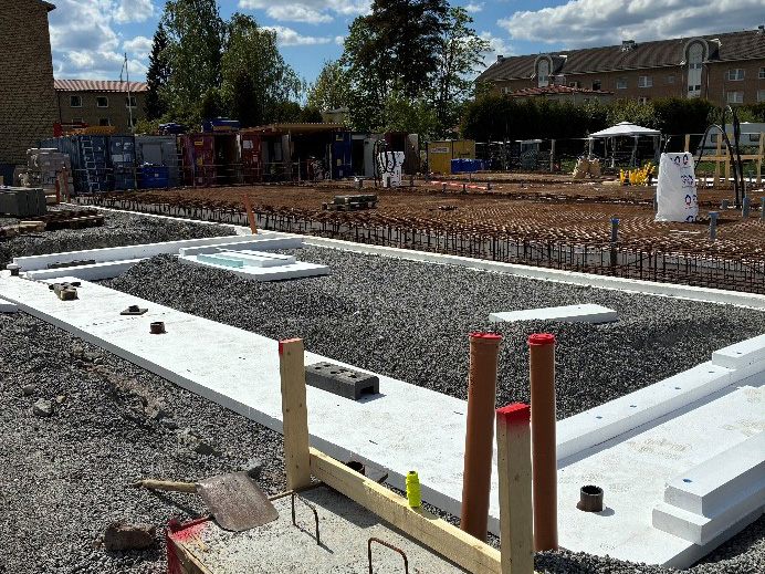En byggarbetsplats med vit isolering, grus och armeringsjärn. "VARNING, BYGGARBETE" står det på ett område i bakgrunden.