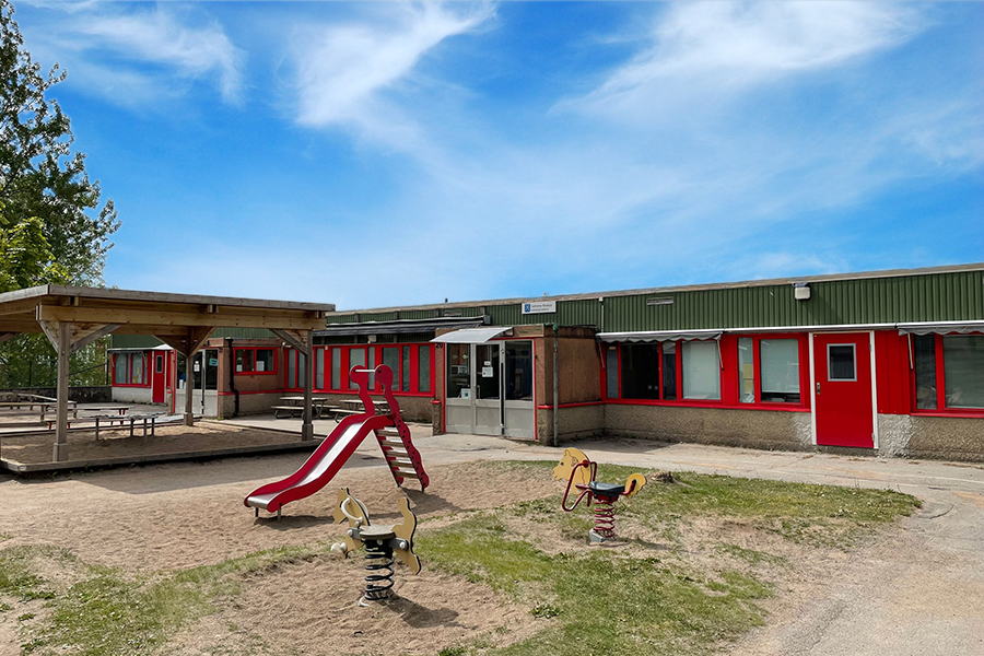 Vallmons förskola. En stenbyggnad med röda detaljer kring fönster och dörrar och en lekpark utanför entrén. 