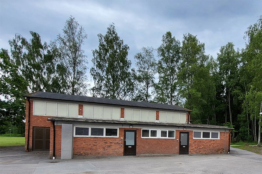 Bilden föreställer en låg byggnad med röd tegelvägg och ett lutande tak. Byggnaden har även en överdel med förtäckta fönster. Det finns flera fönster och två entrédörrar med mörka karmar. Byggnaden står på en asfalterad yta med träd i bakgrunden.