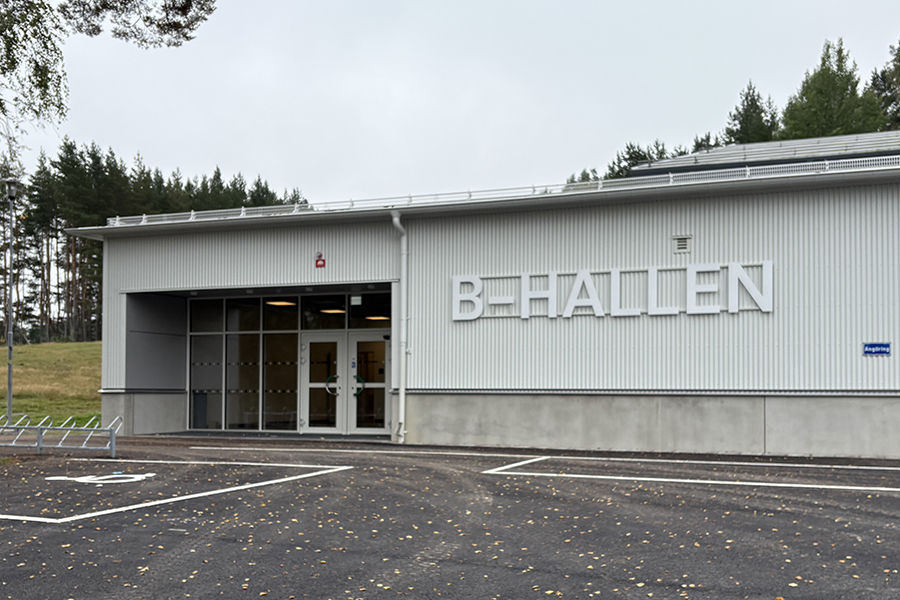 En bild på en entré på en byggnad med ljus fasad. Till vänster om entrén står det b-hallen med versaler.