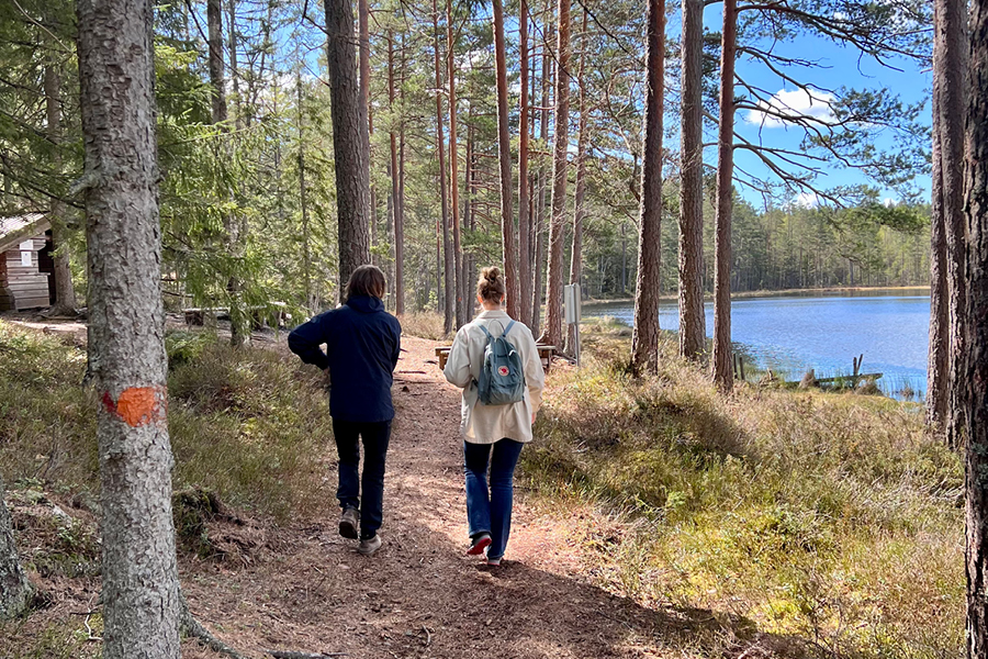 Två personer som går på en vandringsled i skogen med en liten sjö bredvid.