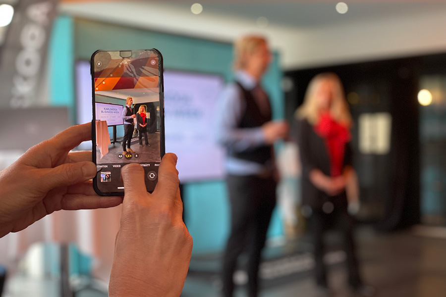 En bild på en telefon med suddig bakgrund där en kvinna och man inviger Business Week