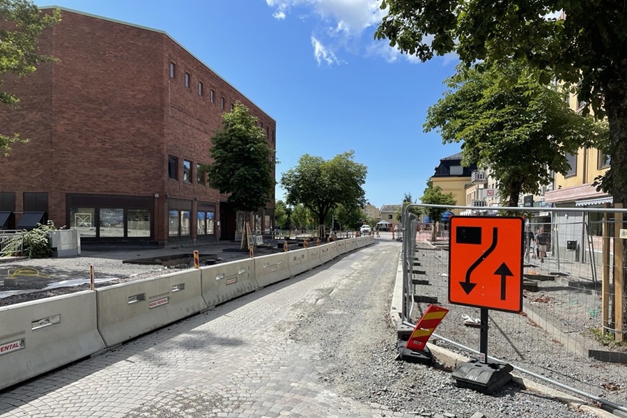 Pågående ombyggnation av en gata i centrala Karlskoga. Gatan är avspärrad med betongbarriärer och stängsel, och en orange skylt visar tillfällig trafikomledning. Träd och byggnader omger arbetsområdet.