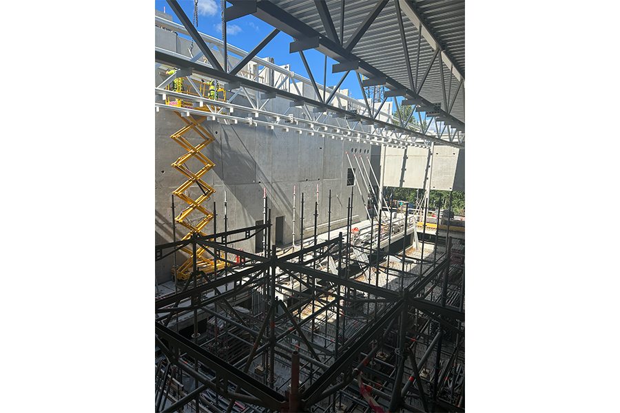 Byggarbetsplats med stålkonstruktion och grå betongväggar. En gul saxlyft och lite vegetation syns i bakgrunden.