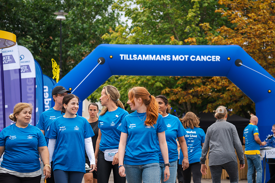 Deltagare i blå Stafett för Livet-tröjor promenerar genom en start- och målportal med texten ”TILLSAMMANS MOT CANCER”. Evenemanget äger rum utomhus i en grönskande parkmiljö, där flera personer rör sig längs banan under arrangemanget.