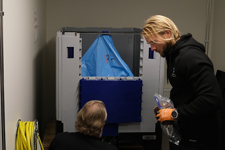 Två personer tittar på en stor blå plastpåse i en grå låda. "OL/EP" syns på påsen.