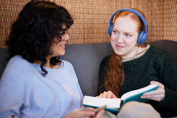Två personer sitter på en soffa och pratar. En kvinna bär blåa hörlurar och har en bok i handen.
