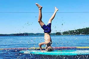 En pojke i badbyxor står på huvudet på en paddelbräda i en sjö med en simlina bakom. Det är soligt och vid horisonten finns en skog.
