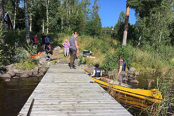 Människor som står runt och på en brygga i vattnet med en kanot bredvid.