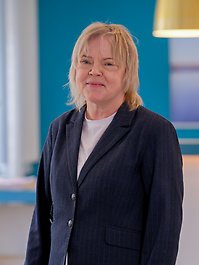 En person med blont hår bär en mörkblå kavaj med vita knappar. Personen ser rakt in i kameran mot en blå bakgrund.