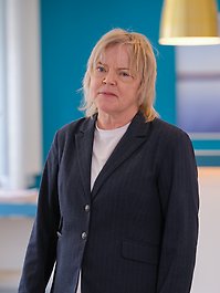 En person med blont hår bär en blårandig kavaj och ser rakt in i kameran. Bakgrunden är en tealfärgad vägg.