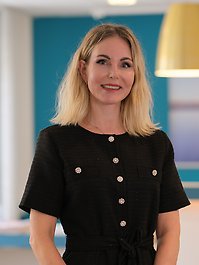 En person med blont hår bär en svart klänning med dekorativa knappar och har armarna i kors.