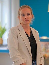 En person i ljusbeige kavaj och svart topp står framför en turkos vägg. Hon har blont hår och ser rakt in i kameran.